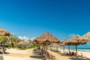 Imagem mostra uma praia de areia clara com várias barracas de palha alinhadas e espreguiçadeiras voltadas para o mar azul, cenário típico do Dom Pedro Laguna, referência entre resorts no Brasil