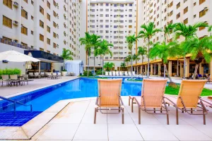 Imagem mostra a área externa do Boulevard Suíte Hotel, com piscina de águas azuis cercada por espreguiçadeiras de palha, mesas com guarda-sóis e palmeiras tropicais, em frente a um edifício de vários andares. O cenário remete ao lazer e conforto característicos dos melhores resorts no Brasil, proporcionando uma experiência completa em Caldas Novas.
