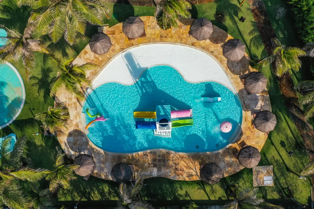 Vista aérea da piscina do Dom Pedro Laguna, com formato orgânico e brinquedos aquáticos coloridos em um espaço infantil. A piscina está cercada por quiosques com cobertura de palha, espreguiçadeiras e coqueiros tropicais, compondo uma área de lazer ideal para famílias. Essa infraestrutura de resort à beira-mar reflete o padrão dos empreendimentos de multipropriedade no Brasil, que oferecem conforto, diversão e férias planejadas com alto nível de qualidade.